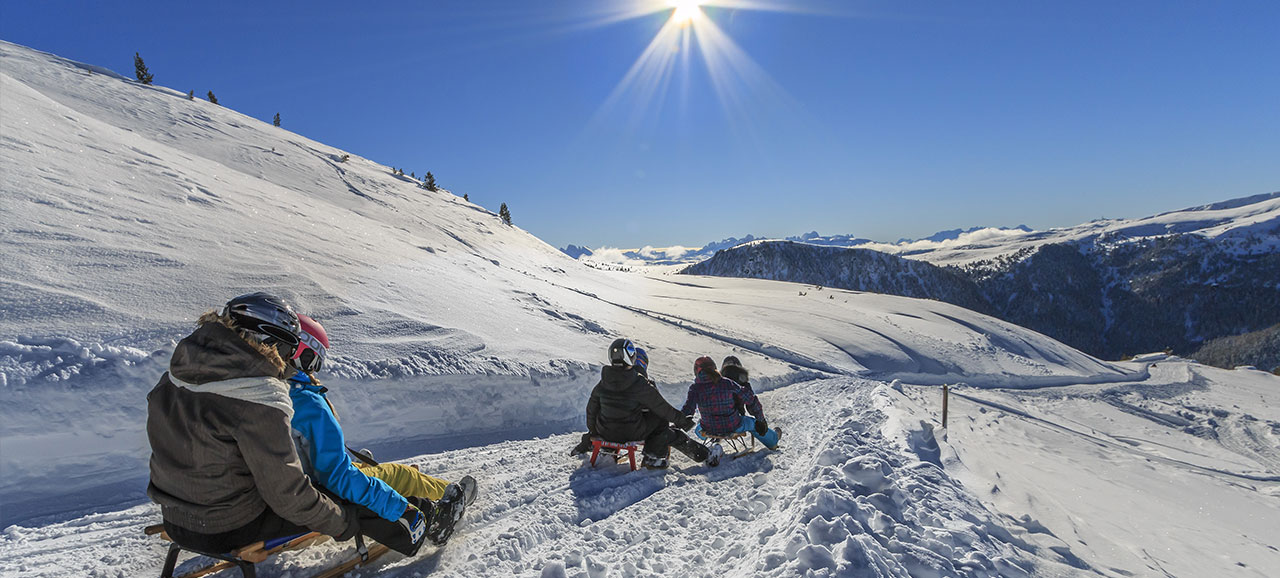 People tobogganing in San Martino (Val Sarentino) in winter with snowy mountains at the back and beautiful sunshine