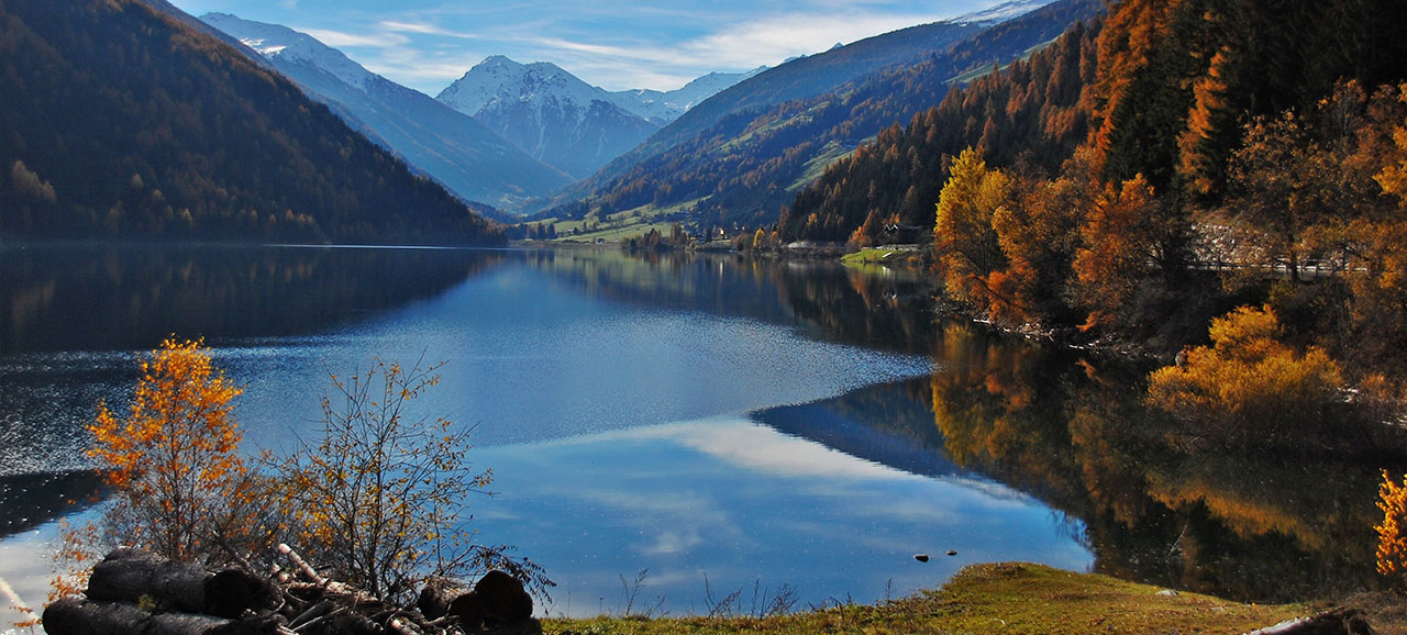 Lago di Zoccolo in autumn, surrounded by autumnal trees and with snowy mountains at the back