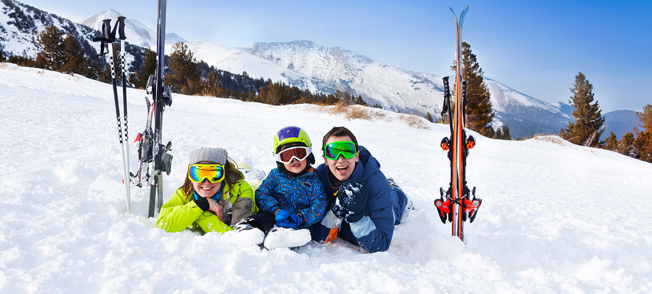 Familie mit Skifahrer Ausrüstung, Helm und Skibrille liegt im Schnee und lächelt in die Kamera