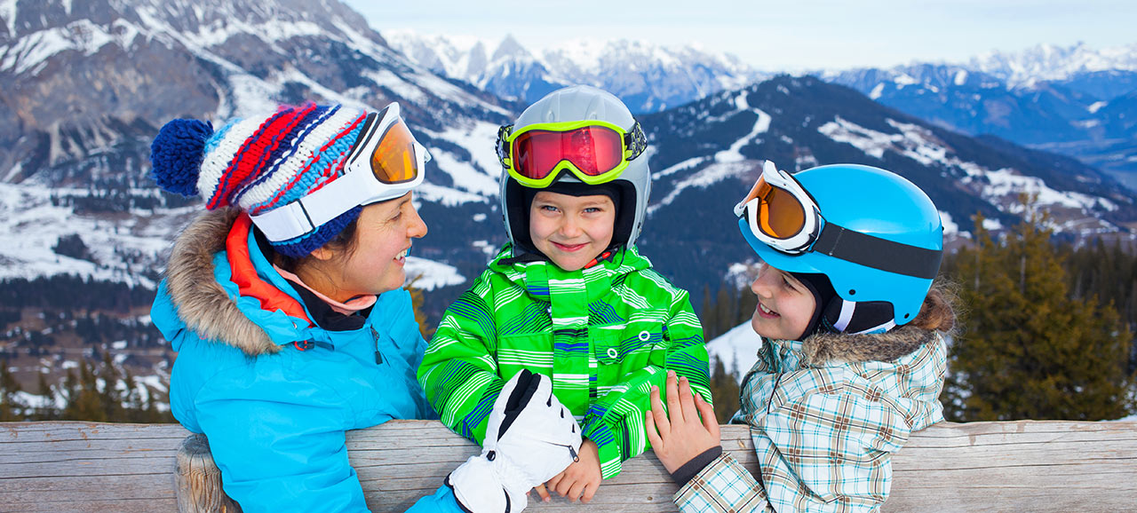 Mother and two children in full skiers equipment at a wooden fence smiling to each other
