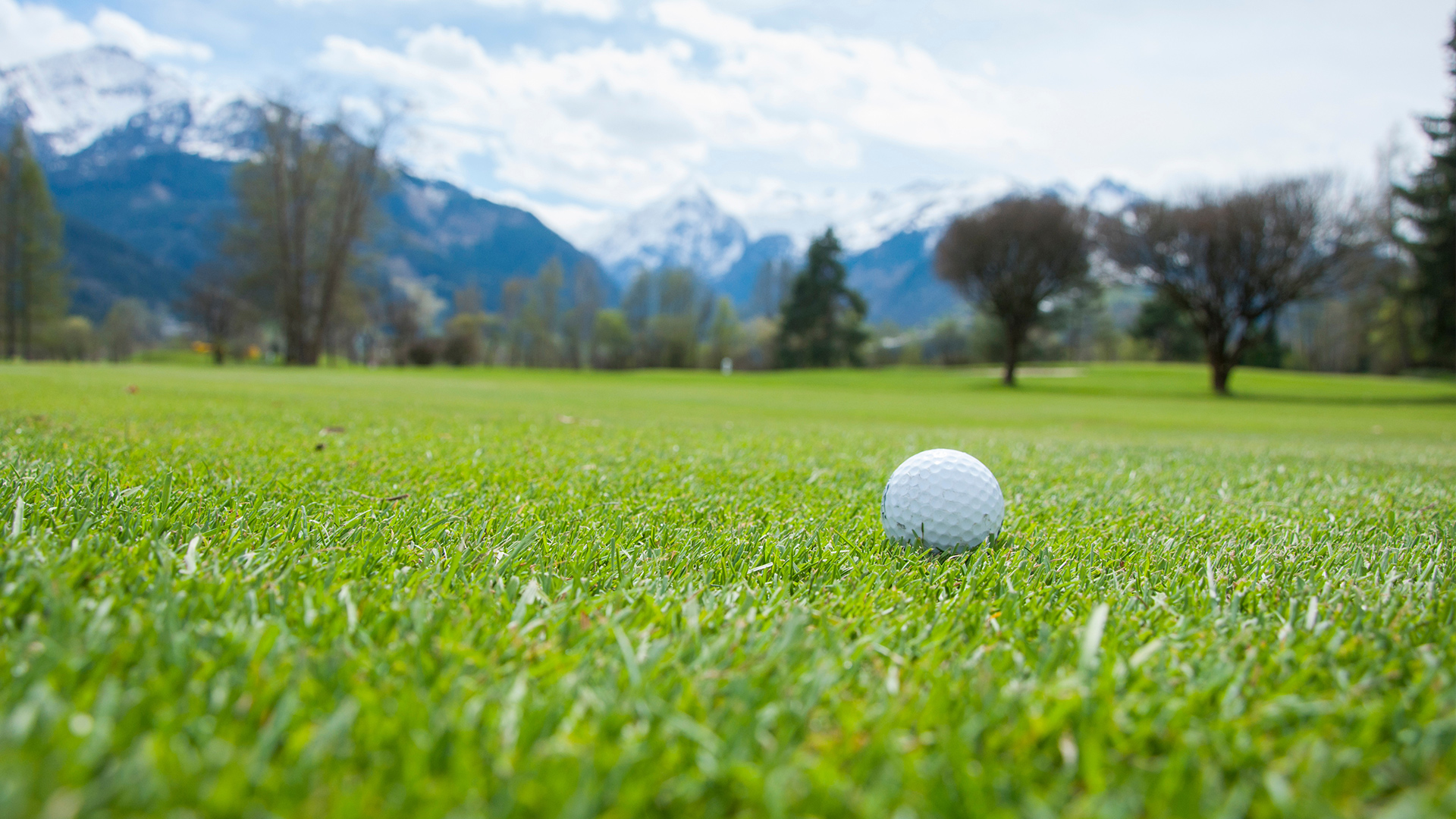 Golfball auf grüner Wiese mit verschneitem Bergpanorama im Hintergrund.
