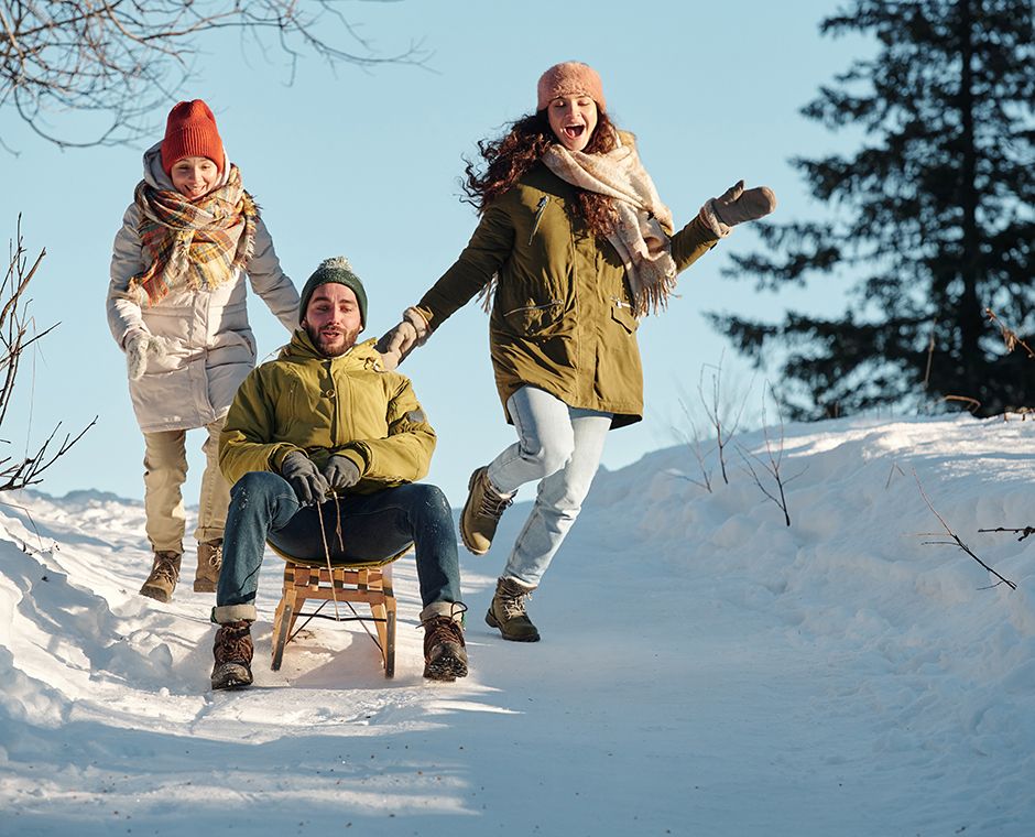Two cheerful girls in winter clothing running behind a youngster on a sled sliding down a hill between snowdrifts in the forest, having fun on a winter day