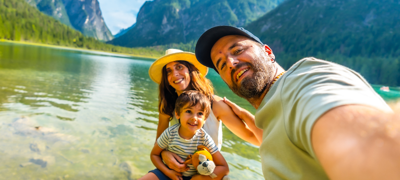 Famiglia felice scatta un selfie al lago tra le montagne con il bimbo in braccio e peluche