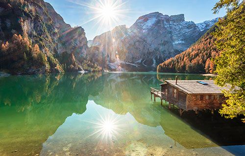 Lago alpino con baita in legno, sole e montagne sullo sfondo