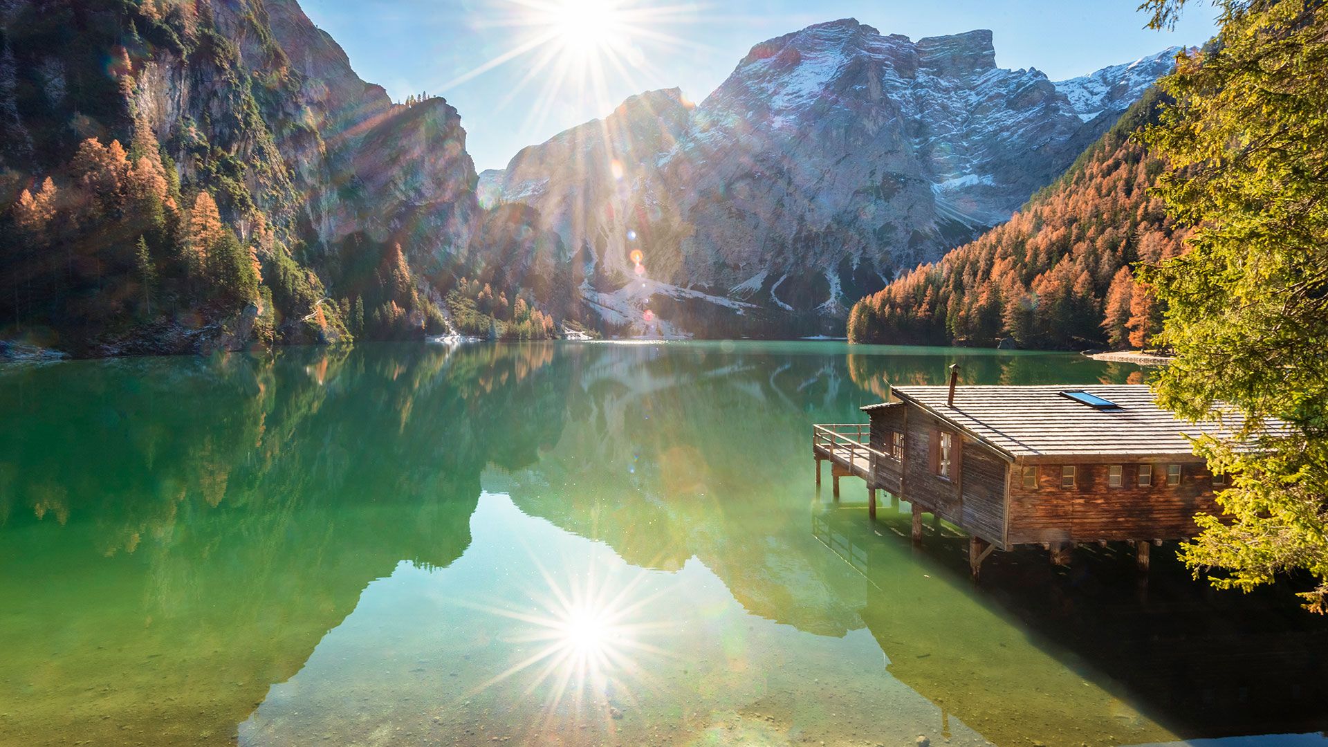 Lago alpino con baita in legno, sole e montagne sullo sfondo
