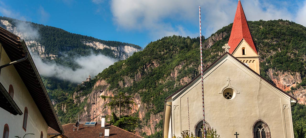 Die zentrale Kirche von Andrian mit dem typischen Glockenturn, gedeckt mit roten Schindeln und den Bergen im Hintergrund