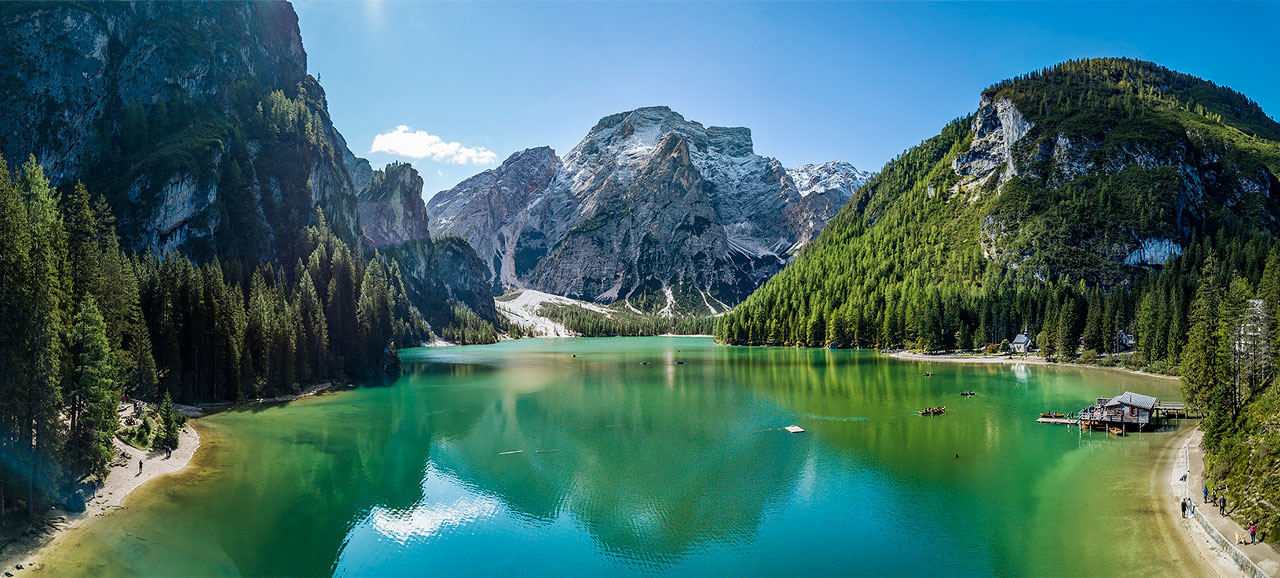 Panoramabild vom Pragser See im Sommer, mit Schiffen im kristallgrünem Wasser und Pfahlhaus aus Holz im Vordergrund