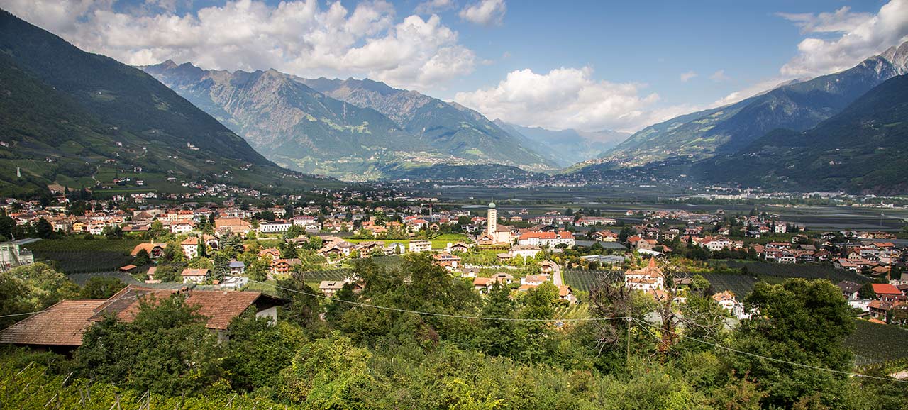 Vista panoramica di Lana con la chiesa di Santa Croce al centro, illuminata dal sole con un cielo parzialmente nuvoloso