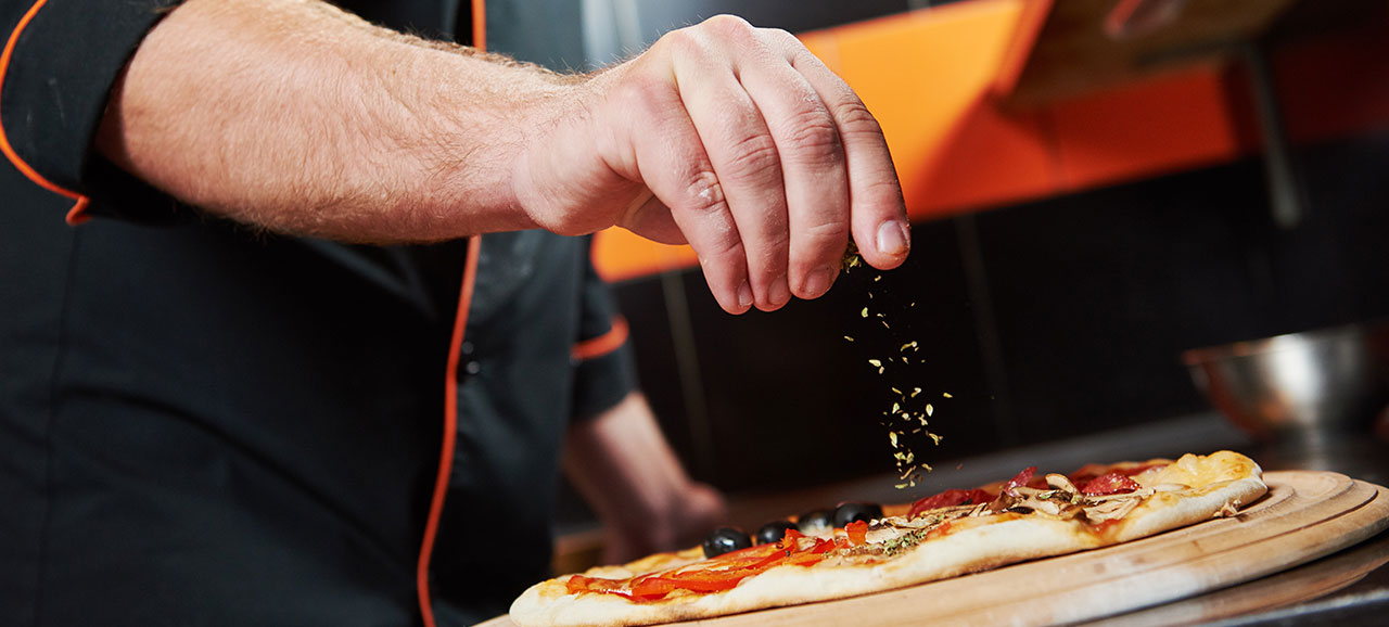 Hand eines Pizzabäckers, der eine Pizza mit Oregano bestreut