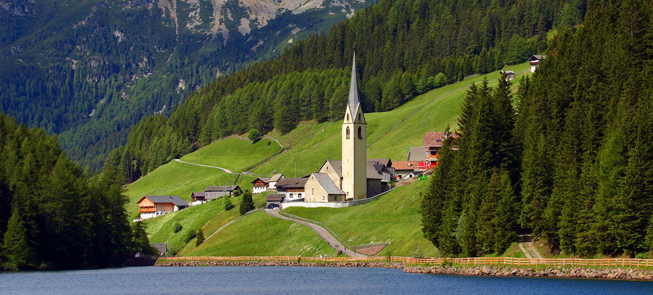 Valdurna Lake in summer with the church and village centre of Valdurna at the back, surrounded by woods and mountains