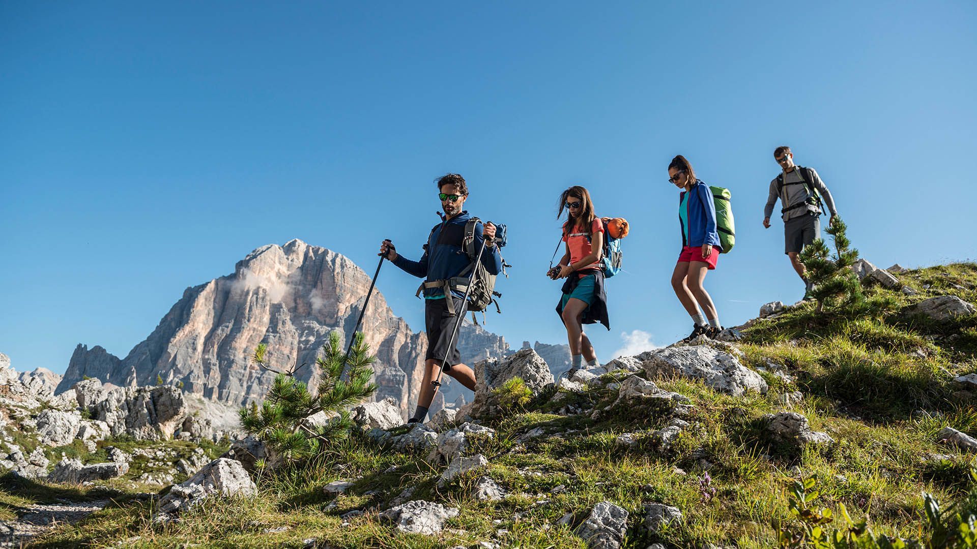 A group of hikers walk among the rocks with a view of the Dolomites on a summer day.