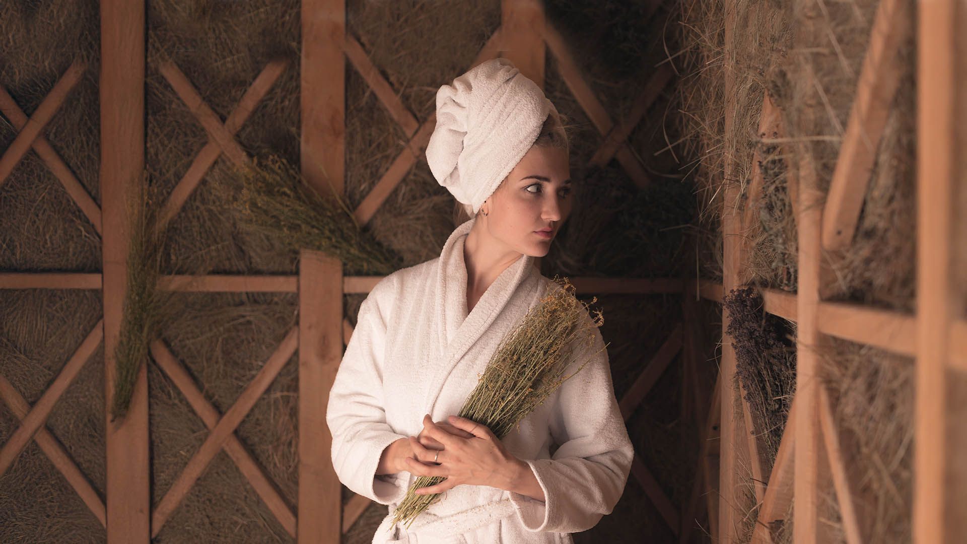 Woman in white bathrobe holding medicinal herbs, inside a hay wellness room