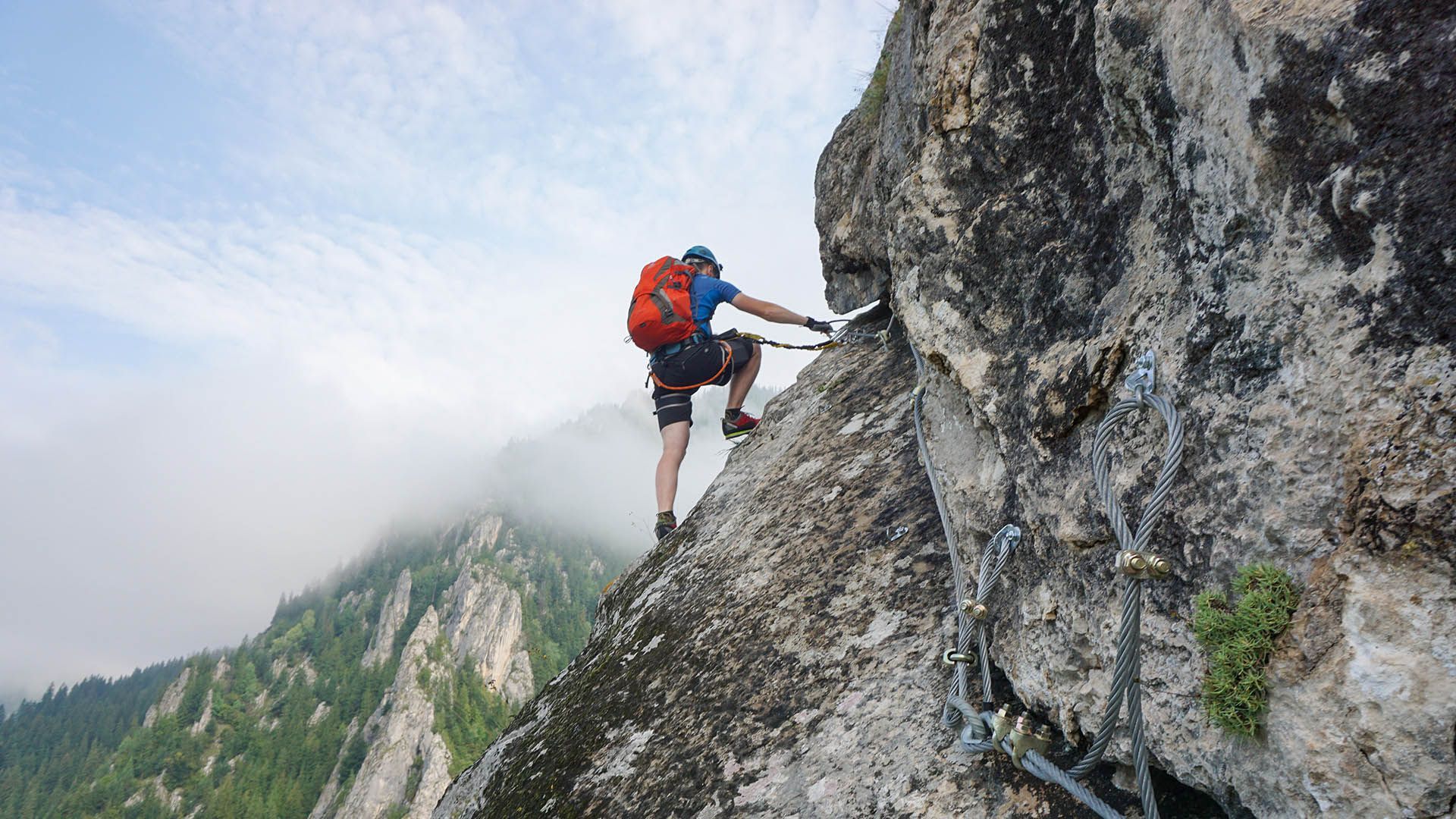 A mountaineer climbs a via ferrata secured with carabiners and ropes.