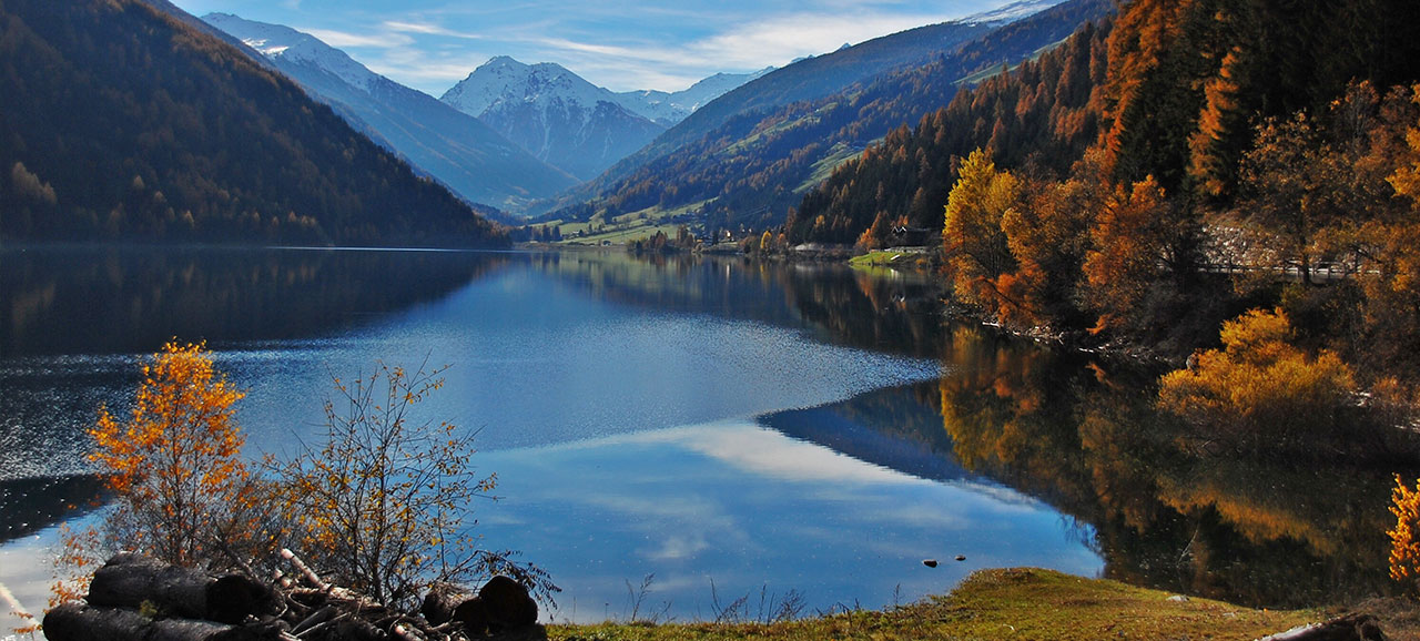 Zoggler Stausee im Herbst, von Bäumen mit Herbstfarben umgeben und mit verschneiten Bergen im Hintergrund