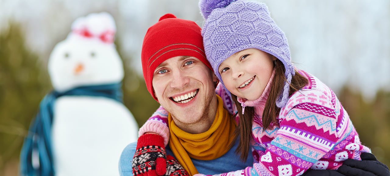 Padre e bambina con cappello di lana, abbracciati, con un pupazzo di neve sullo sfondo