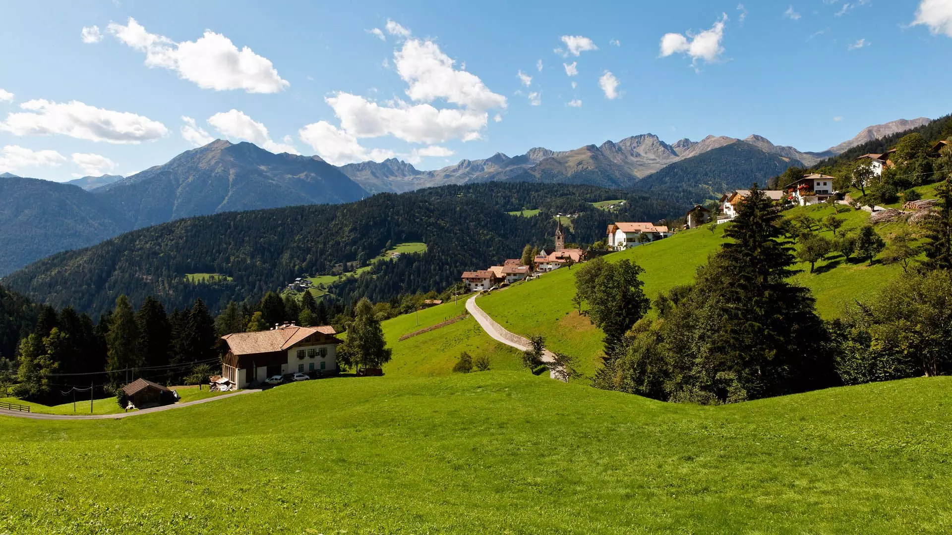 Blick auf Laurein zwischen grünen Wiesen und Bergen, mit Kirche und Alpenhäusern unter dem blauen Himmel